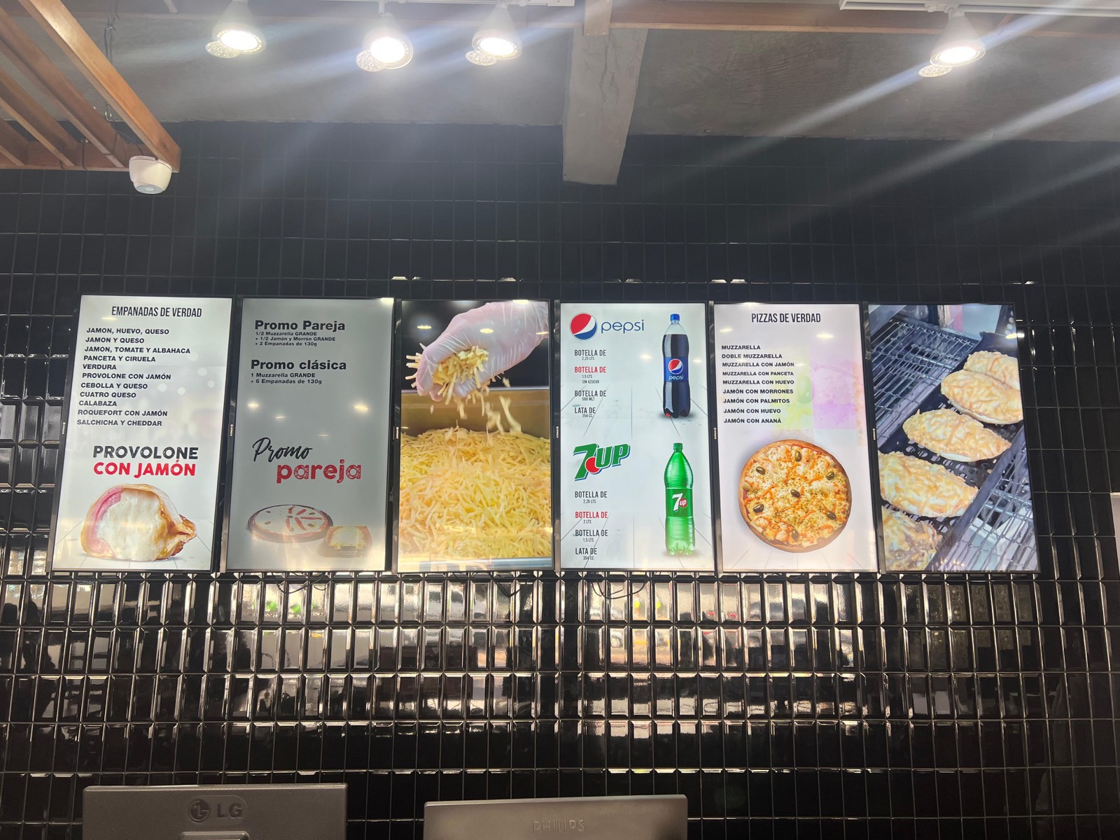 Seis pantallas sobre el mostrador de una pizzería mostrando menús, promociones y precios